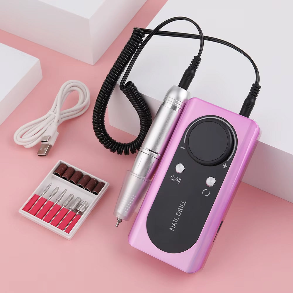 Ponceuse à ongles 45 000 RPM rechargeable en finition métallique rose-violet, avec pièce à main en aluminium, câble spiralé et kit de forets et embouts abrasifs, posée sur une surface rose.